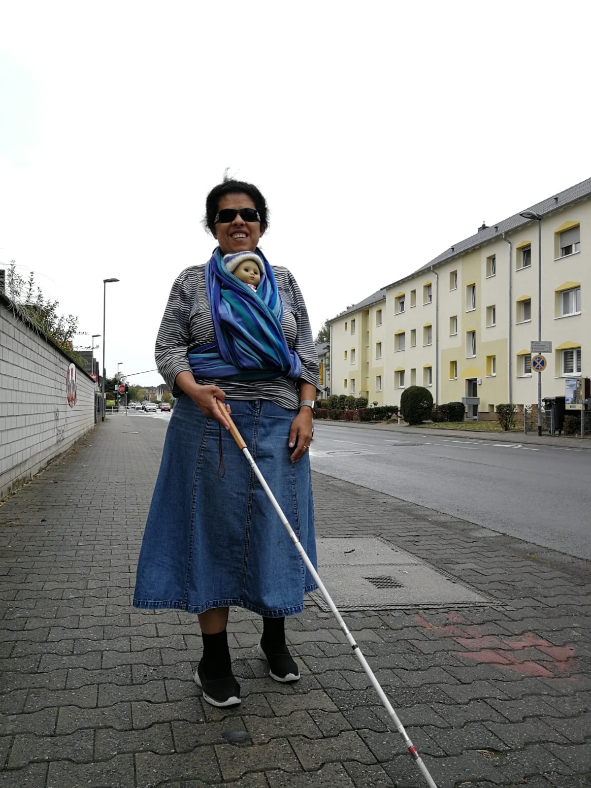 Lydia läuft mit Puppe im Tragetuch und Blindenstock eine Straße entlang.