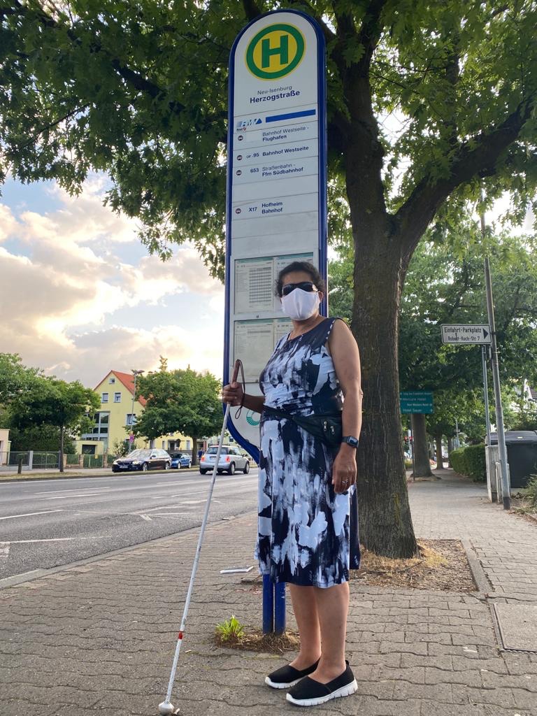 Lydia steht mit Maske und Blindenlangstock an einer Bushaltestelle.