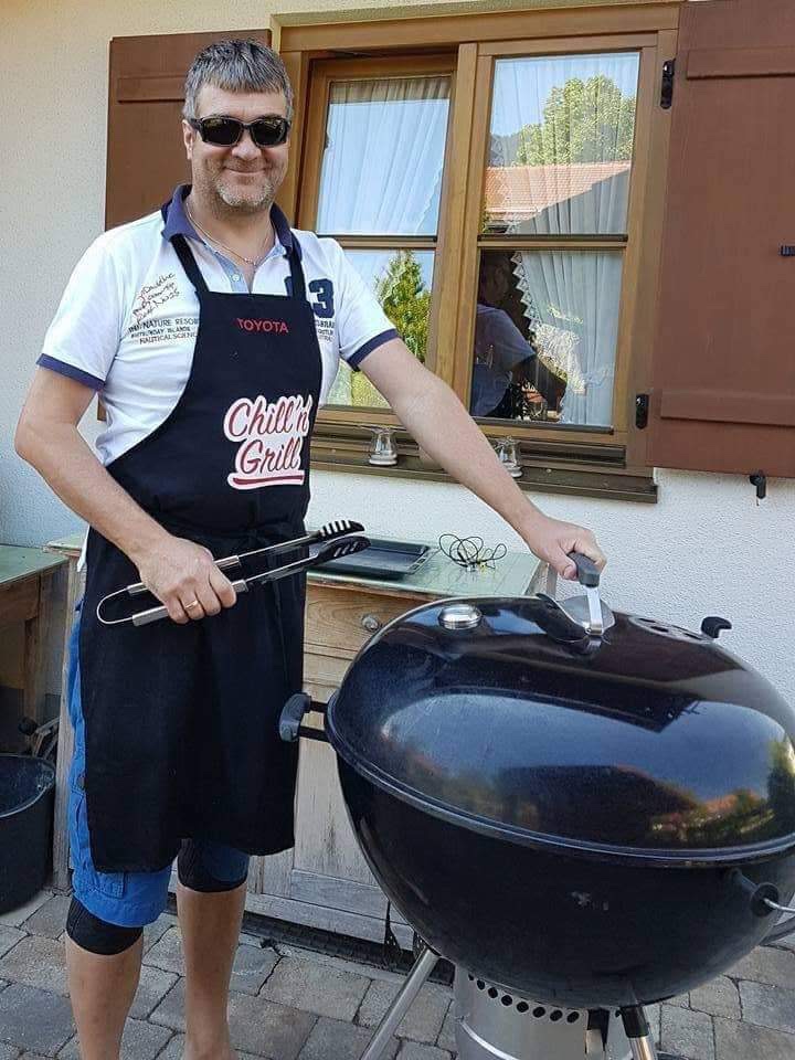 Marcus mit Grillgabel in der rechten Hand und die linke am Griff des Deckel eines Kugelgrills. Schürze mit der Aufschrift "Chill 'n Grill"