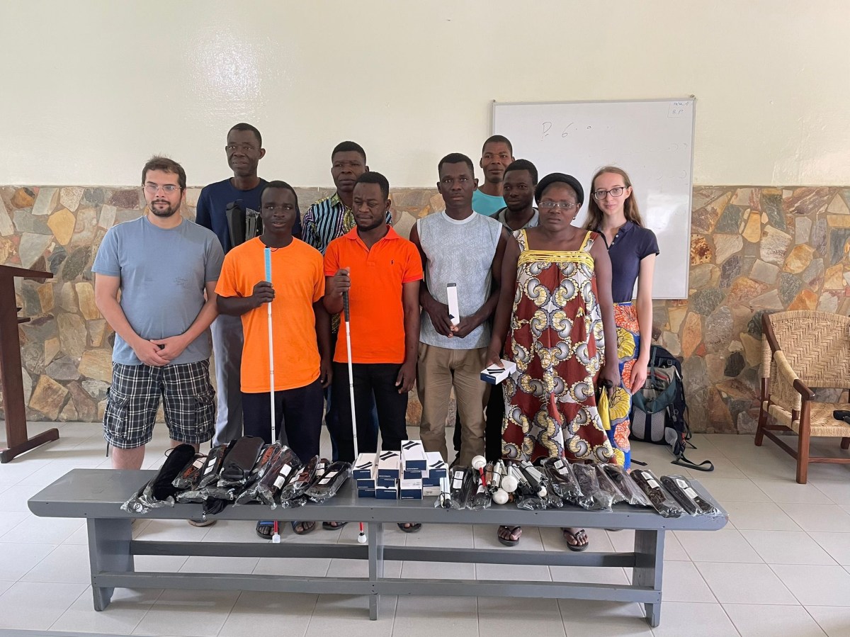 acht. Lehrer:innen des. Blindenzentrums sowie. Jasmin und ihren Mann. Vor ihnen auf. einer Bank sind die gespendeten Langstöcke. und Lupen aufgestapelt. weitere werden von den. Lehrer:innen in der Hand. gehalten.