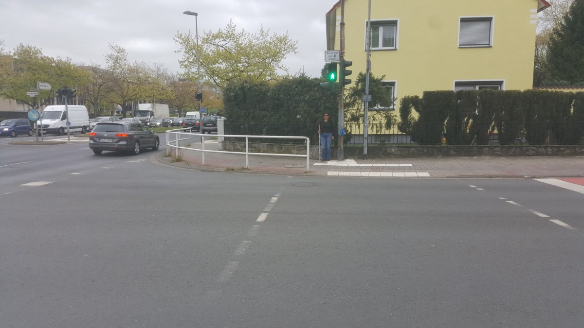 Lydia steht mit Blindenlangstock an der Ecke einer stark befahrenen Straße.
