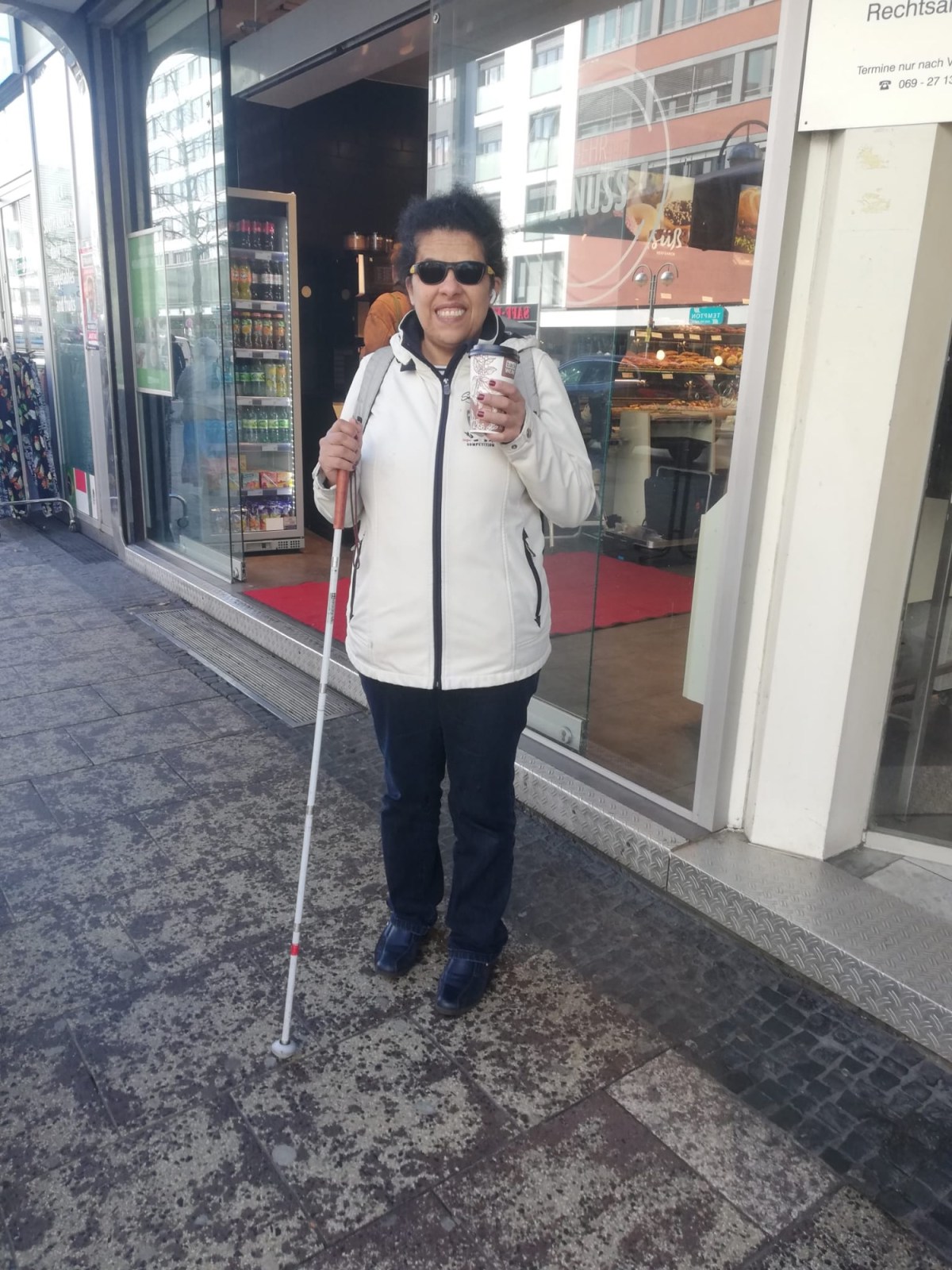 Lydia steht mit Blinden Langstock neben einem Bäcker. In der Hand hält sie einen Kaffeebecher